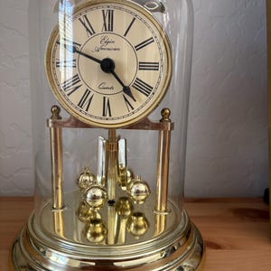 May include: A gold-toned Elgin American quartz clock with a cream-colored face and black Roman numerals. The clock is encased in a clear glass dome and features a pendulum with gold spheres. The clock's base is also gold.