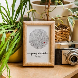May include: A framed print with a map of a city in a circle. The map is in black and white with a red heart marking a location. The text below the map reads "georgia &hearts; bob 51 Booth Avenue 14.08.2023".