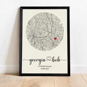 May include: A black framed print with a map of a city in a circle with a red heart in the center. The text below the map reads "georgia &hearts; bob" and "51 Booth Avenue 14.08.2023".