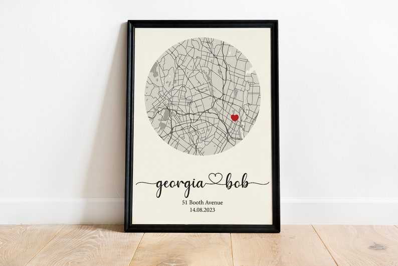 May include: A black framed print with a map of a city in a circle with a red heart in the center. The text below the map reads "georgia &hearts; bob" and "51 Booth Avenue 14.08.2023".