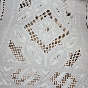 Könnte beinhalten: Weißer bestickter Tischläufer mit geometrischen und floralen Mustern. Das Design weist Lochmuster und Vollstich auf, wodurch ein texturierter Effekt entsteht. Die Kanten des Läufers sind mit einem gewellten Rand versehen.