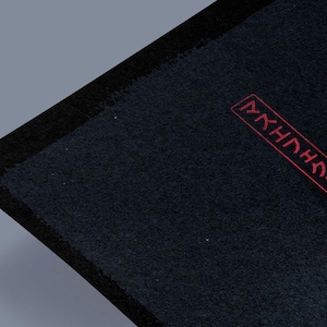 May include: A dark blue rectangular textile with a black border. The upper right corner features red Japanese characters within a rectangular frame. The textile has a textured surface and is set against a gray background.
