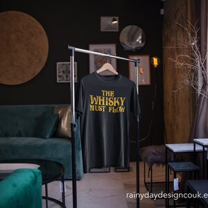 May include: Black t-shirt with the text "The Whisky Must Flow" in gold lettering. The shirt is hanging on a black clothing rack in a room with a green sofa and a wooden floor.