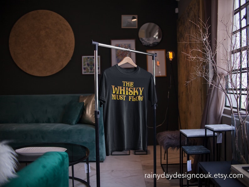 May include: Black t-shirt with the text "The Whisky Must Flow" in gold lettering. The shirt is hanging on a black clothing rack in a room with a green sofa and a wooden floor.