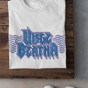 May include: White t-shirt with a blue graphic that says "WISTLE DEATH" with wavy lines on either side. The shirt is folded and laid on a wooden surface. There is a small logo in the top right corner that says "Rainy Day Design Co." with a cloud and raindrops.