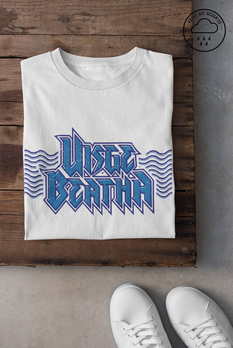 May include: White t-shirt with a blue graphic that says "WISTLE DEATH" with wavy lines on either side. The shirt is folded and laid on a wooden surface. There is a small logo in the top right corner that says "Rainy Day Design Co." with a cloud and raindrops.