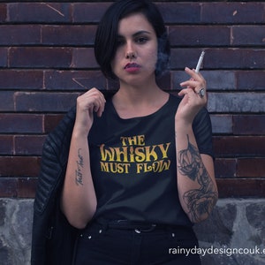 May include: A woman wearing a black t-shirt with the text "The Whisky Must Flow" in yellow lettering. She is standing in front of a brick wall and has a tattoo on her arm.