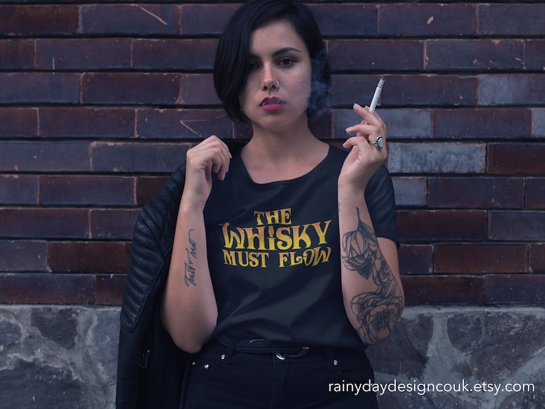 May include: A woman wearing a black t-shirt with the text "The Whisky Must Flow" in yellow lettering. She is standing in front of a brick wall and has a tattoo on her arm.