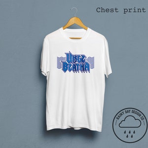 May include: White t-shirt with a blue graphic print of the text "WISTERIA" with wavy lines above and below the text. The text is in a bold, stylized font. The t-shirt is hanging on a wooden hanger against a light blue background. The logo "RAINY DAY DESIGN CO." is in the lower right corner of the image.