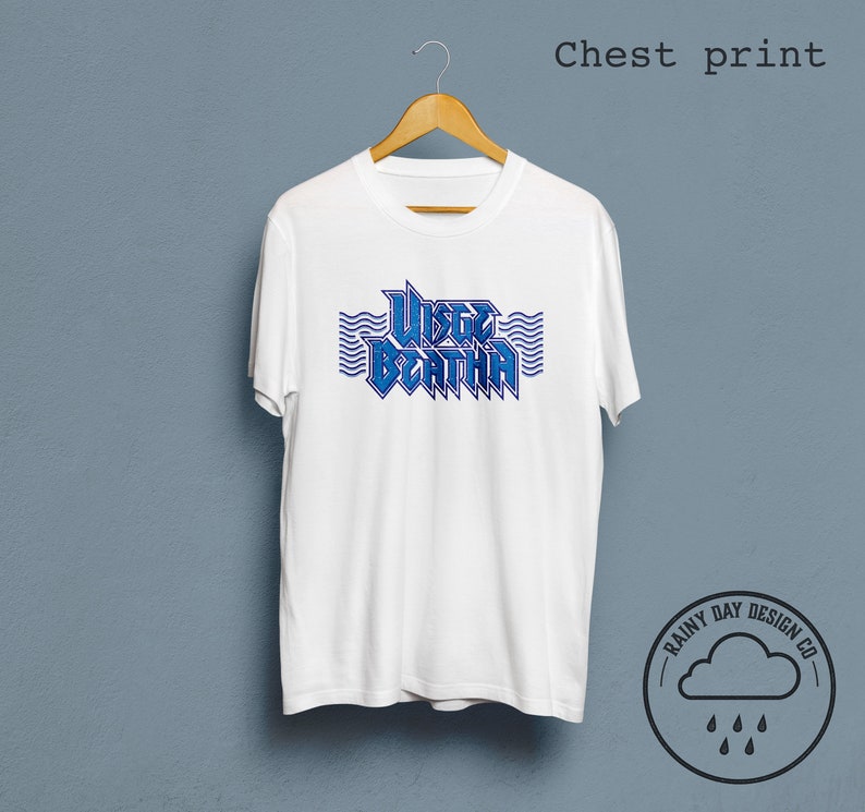 May include: White t-shirt with a blue graphic print of the text "WISTERIA" with wavy lines above and below the text. The text is in a bold, stylized font. The t-shirt is hanging on a wooden hanger against a light blue background. The logo "RAINY DAY DESIGN CO." is in the lower right corner of the image.