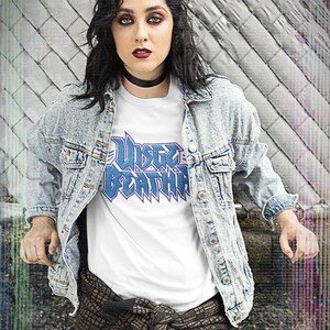 May include: A woman wearing a white t-shirt with a blue graphic that says "W&uuml;sten Bestia" and a light blue denim jacket. She is wearing a brown and black patterned scarf tied around her waist and blue jeans.