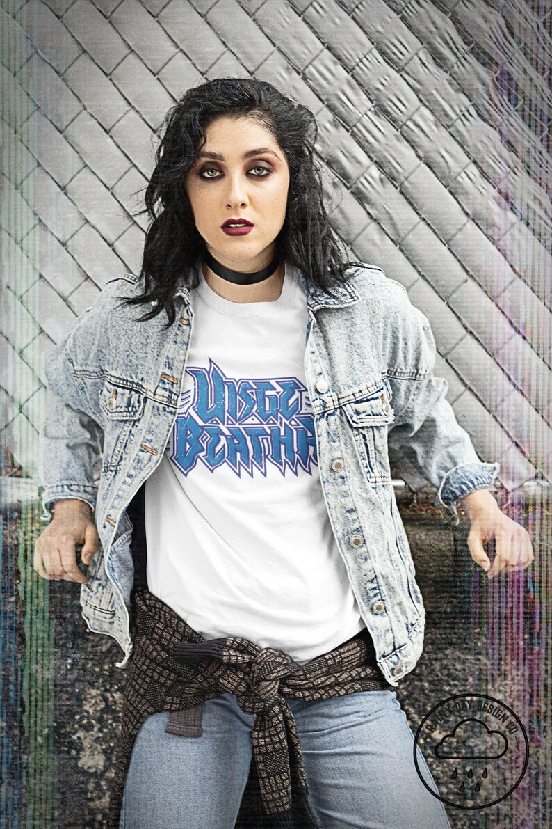 May include: A woman wearing a white t-shirt with a blue graphic that says "W&uuml;sten Bestia" and a light blue denim jacket. She is wearing a brown and black patterned scarf tied around her waist and blue jeans.