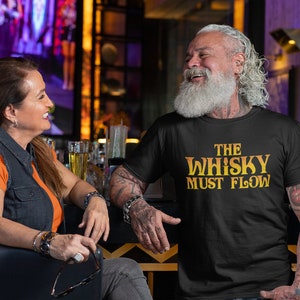 May include: A man with a long white beard is wearing a black t-shirt with the text "The Whisky Must Flow" in yellow. He is smiling and looking to the right. He has tattoos on his arms.