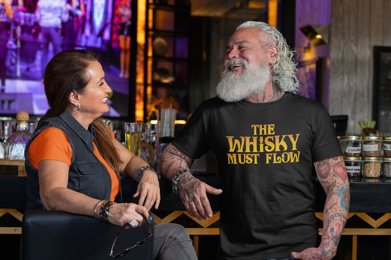 May include: A man with a long white beard is wearing a black t-shirt with the text "The Whisky Must Flow" in yellow. He is smiling and looking to the right. He has tattoos on his arms.