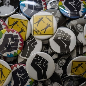 May include: A collection of round buttons with various designs. Some buttons feature a black fist raised in the air, while others have a yellow warning sign with the text "DANGER POLICE IN AREA".