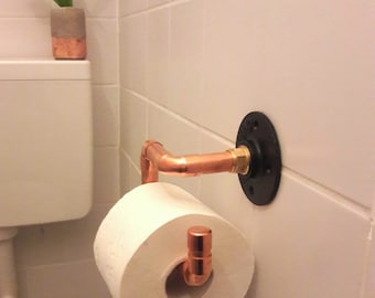 Op de afbeelding: Toiletpapierhouder van koperen pijp, gemonteerd op een witte betegelde muur. De houder heeft een zwarte flens en een koperen arm met een koperen haak. Een rol wit toiletpapier zit op de haak.