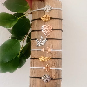 Può includere: Una collezione di otto braccialetti con ciondoli diversi, tra cui un albero, una foglia, un cuore, un uccello, un'ancora, un'ala, una croce e un design circolare. I braccialetti sono esposti su una superficie di legno.
