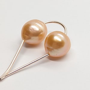 Peut inclure: Une paire de boucles d'oreilles avec deux perles de couleur pêche. Chaque perle est montée sur un fil d'argent délicat, avec un accent argenté incurvé les reliant. Les boucles d'oreilles sont posées sur un fond blanc.