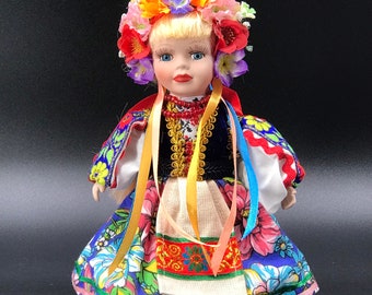 May include: A porcelain doll wearing a traditional folk costume with a colorful floral wreath. The dress is blue with floral patterns and yellow, red, and white ribbons.