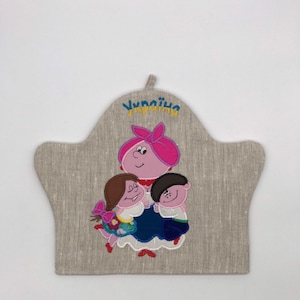 Pode incluir: Um pegador de panela de linho bege com um desenho bordado de uma mulher e duas crianças com roupas tradicionais ucranianas. O texto "Україна" está bordado acima das figuras.