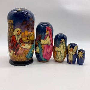 May include: A set of five nesting dolls, each painted with religious figures and scenes. The largest doll depicts a nativity scene with a star, while the others show figures in robes and a star. The dolls are primarily blue, with accents of red, gold, and white.