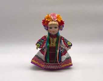 May include: A porcelain doll in traditional Ukrainian attire. The doll has blonde hair, blue eyes, and wears a floral crown. The dress is red, white, and yellow with intricate embroidery and colorful ribbons.