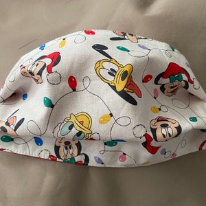 Op de afbeelding: Witte stoffen gezichtsmasker met een herhaald patroon van Mickey Mouse, Minnie Mouse, Pluto en Donald Duck. De karakters worden afgewisseld met kleurrijke kerstverlichting. Het masker heeft een rode binnenvoering.