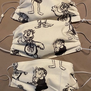 Puede incluir: Tres mascarillas de tela blanca con ilustraciones en blanco y negro de personajes de Los Muppets. Las mascarillas representan a Kermit la rana, Miss Piggy y Fozzie Bear.