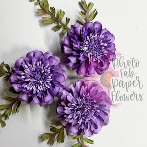 Può includere: Tre fiori di carta viola con petali a strati e fogliame di carta verde. I fiori presentano un centro strutturato e sono disposti a grappolo. Il testo Photo Fab Paper Flowers è stampato in corsivo verso il lato destro della composizione.