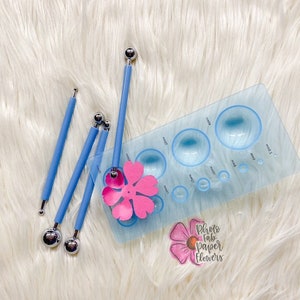 May include: A set of four blue tools with round metal tips and a blue plastic template with multiple circular cutouts. The template has a pink paper flower on it. The text "Photo Fab Paper Flowers" is visible on the template.