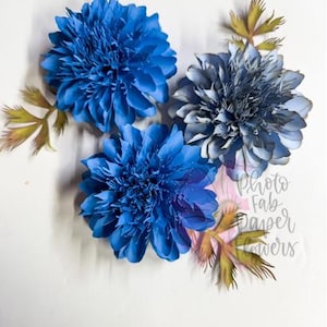 May include: Three handmade paper flowers in shades of blue and gray, with green and yellow accents. The flowers have detailed petal structures and are arranged on a white background. The image includes the text "Photo Fab Paper Flowers".