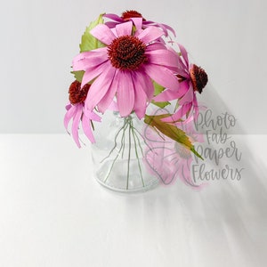K&ouml;nnte beinhalten: Ein Strau&szlig; rosa Papierblumen mit braunen Zentren in einer klaren Glasvase. Die Blumen sind kreisf&ouml;rmig angeordnet und die Vase steht auf einer wei&szlig;en Oberfl&auml;che. Der Text "Photo Fab Paper Flowers" ist in der unteren rechten Ecke des Bildes sichtbar.