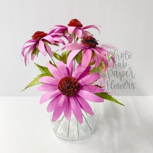 K&ouml;nnte beinhalten: Ein Strau&szlig; rosa Papierblumen in einer durchsichtigen Glasvase. Die Blumen haben eine rote Mitte und sind in einer Vase auf wei&szlig;em Hintergrund angeordnet.