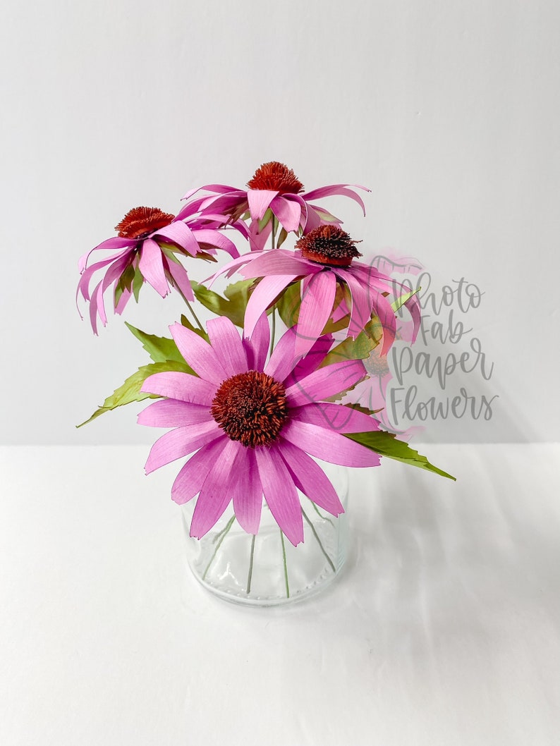 K&ouml;nnte beinhalten: Ein Strau&szlig; rosa Papierblumen in einer durchsichtigen Glasvase. Die Blumen haben eine rote Mitte und sind in einer Vase auf wei&szlig;em Hintergrund angeordnet.