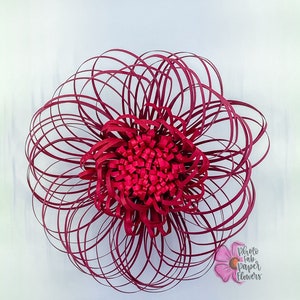 May include: A large, burgundy paper flower with a layered, circular design. The flower is made of many thin strips of paper that are curled and layered to create a 3D effect. The flower is centered in the image and is the only object present.