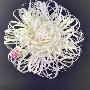 May include: A large white paper flower with a layered, ruffled design. The flower is made from thin strips of paper that are curled and twisted to create a 3D effect. The flower is against a dark gray background.