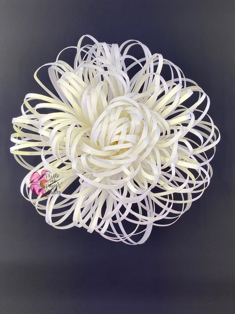 May include: A large white paper flower with a layered, ruffled design. The flower is made from thin strips of paper that are curled and twisted to create a 3D effect. The flower is against a dark gray background.