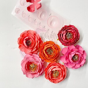 May include: Six paper flowers in various shades of pink and orange. The flowers are arranged in a circle on a white surface. A plastic flower shaping tool is also visible in the image.