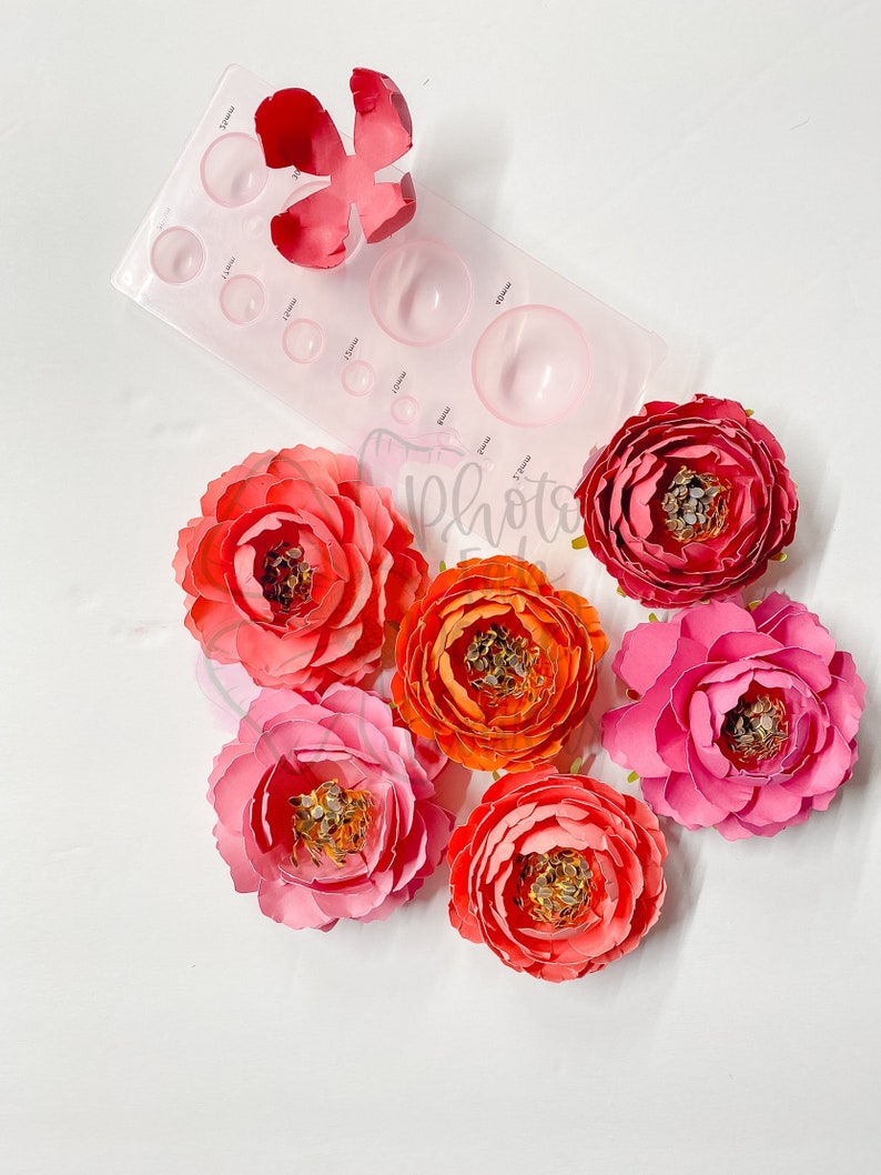 May include: Six paper flowers in various shades of pink and orange. The flowers are arranged in a circle on a white surface. A plastic flower shaping tool is also visible in the image.
