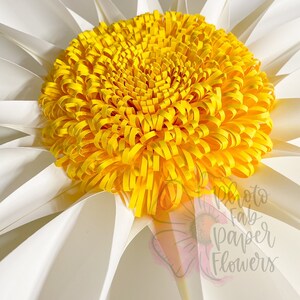 May include: A large white paper daisy with a yellow paper center. The center is made of many curled paper strips. The daisy is a paper flower.