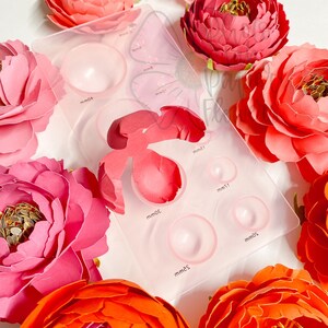 May include: A clear plastic template with eight different sized circles for creating paper flowers. The template is surrounded by several paper flowers in various shades of pink and orange.
