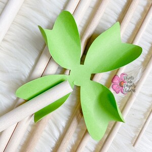 May include: A green paper flower with five petals is attached to a wooden dowel. The flower is on a white fluffy surface with other wooden dowels.