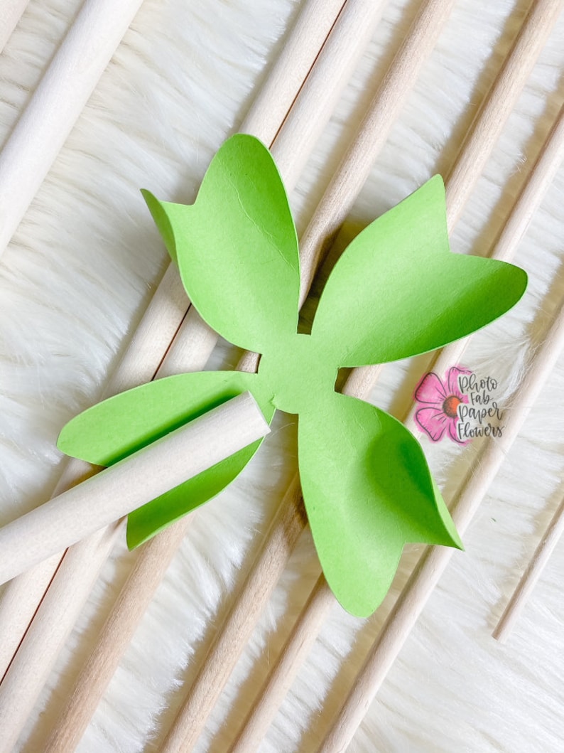 May include: A green paper flower with five petals is attached to a wooden dowel. The flower is on a white fluffy surface with other wooden dowels.