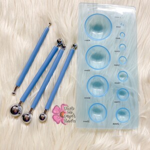 May include: A set of four blue handled tools with round metal tips of varying sizes, used for creating paper flowers. The tools are next to a clear plastic template with nine round indentations of different sizes, labeled with measurements in millimeters.