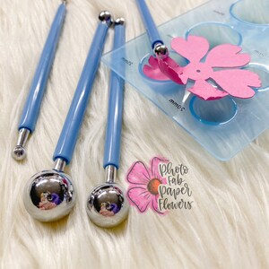 May include: Four blue handled tools with round metal tips, used for creating paper flowers. The tools are on a white fuzzy surface next to a clear plastic template with circles and a pink paper flower.