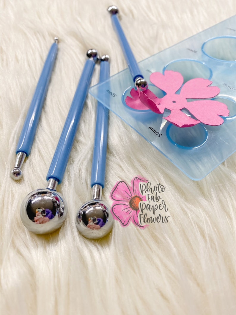May include: Four blue handled tools with round metal tips, used for creating paper flowers. The tools are on a white fuzzy surface next to a clear plastic template with circles and a pink paper flower.