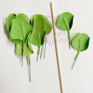May include: A set of green paper leaves with wire stems, perfect for crafting. The leaves are arranged on a white surface next to a wooden stick.