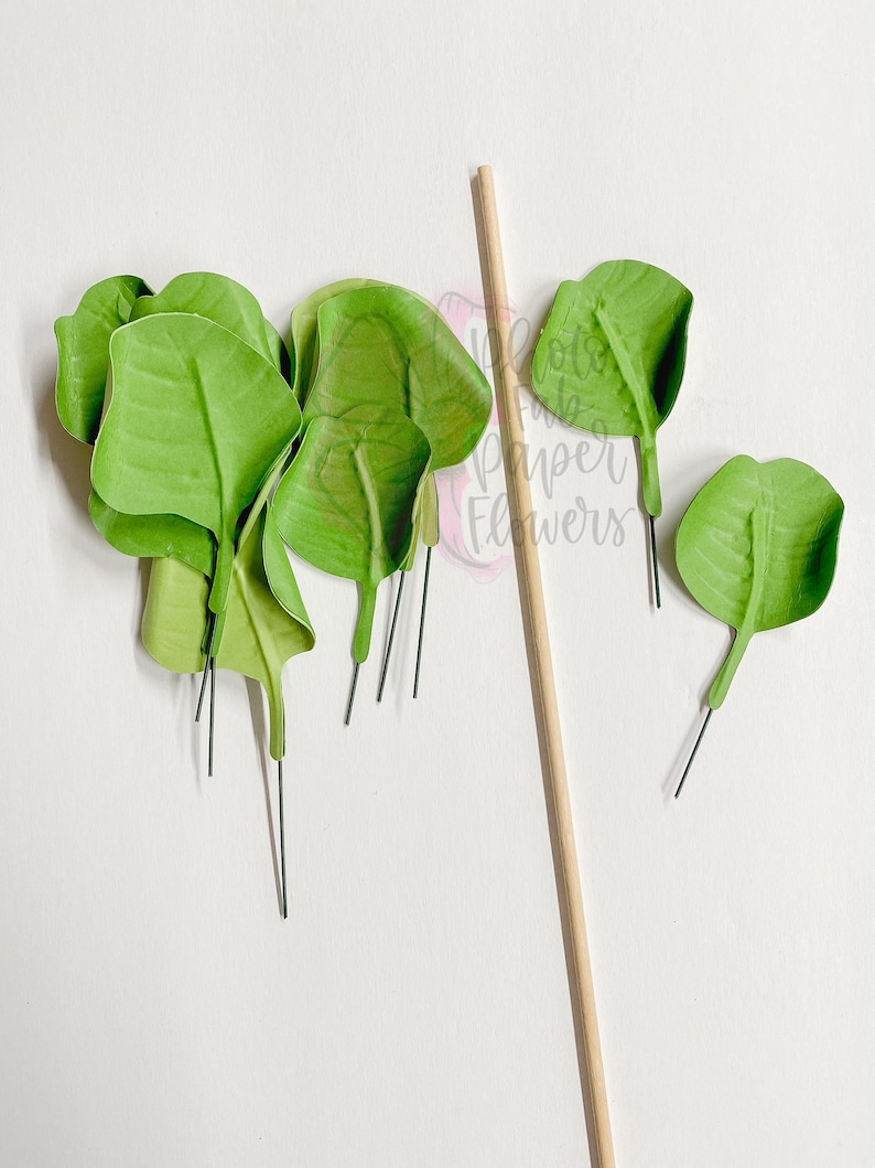 May include: A set of green paper leaves with wire stems, perfect for crafting. The leaves are arranged on a white surface next to a wooden stick.