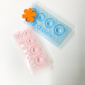 May include: Two plastic circle templates for paper crafting. The blue template has 8 circles ranging in size from 1 cm to 6 cm. The pink template has 10 circles ranging in size from 1 cm to 5 cm.