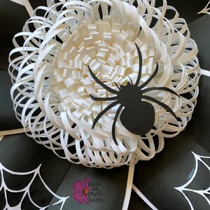 May include: A white paper flower with a black spider on top. The flower is made of many layers of paper, creating a 3D effect. The spider is a simple silhouette with eight legs. The background is black with white spiderwebs.
